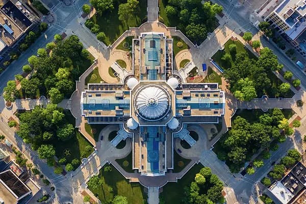 A symmetrical high-angle aerial view of a grand dome-topped capitol building surrounded by manicured lawns, circular plazas, and radial pathways, representing civic architecture and government infrastructure.