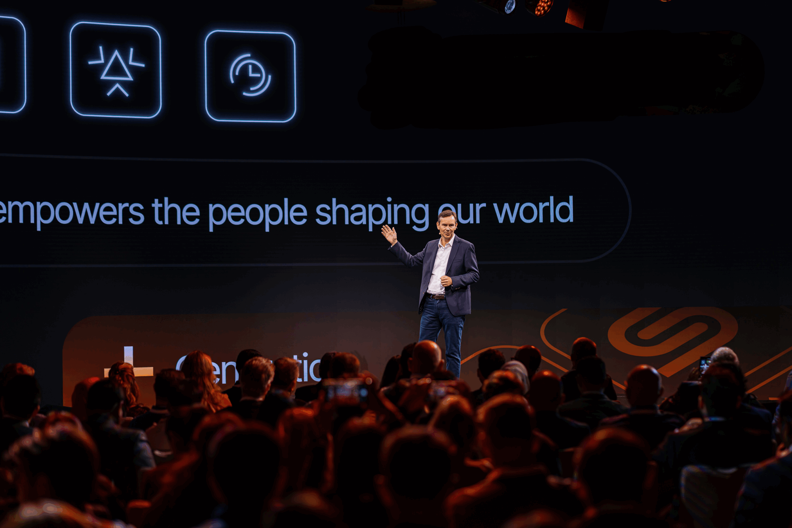 A man in business attire gives a presentation on stage to an audience, with a large screen displaying digital icons and text in the background. A man in business attire gives a presentation on stage to an audience, with a large screen displaying digital icons and text in the background.