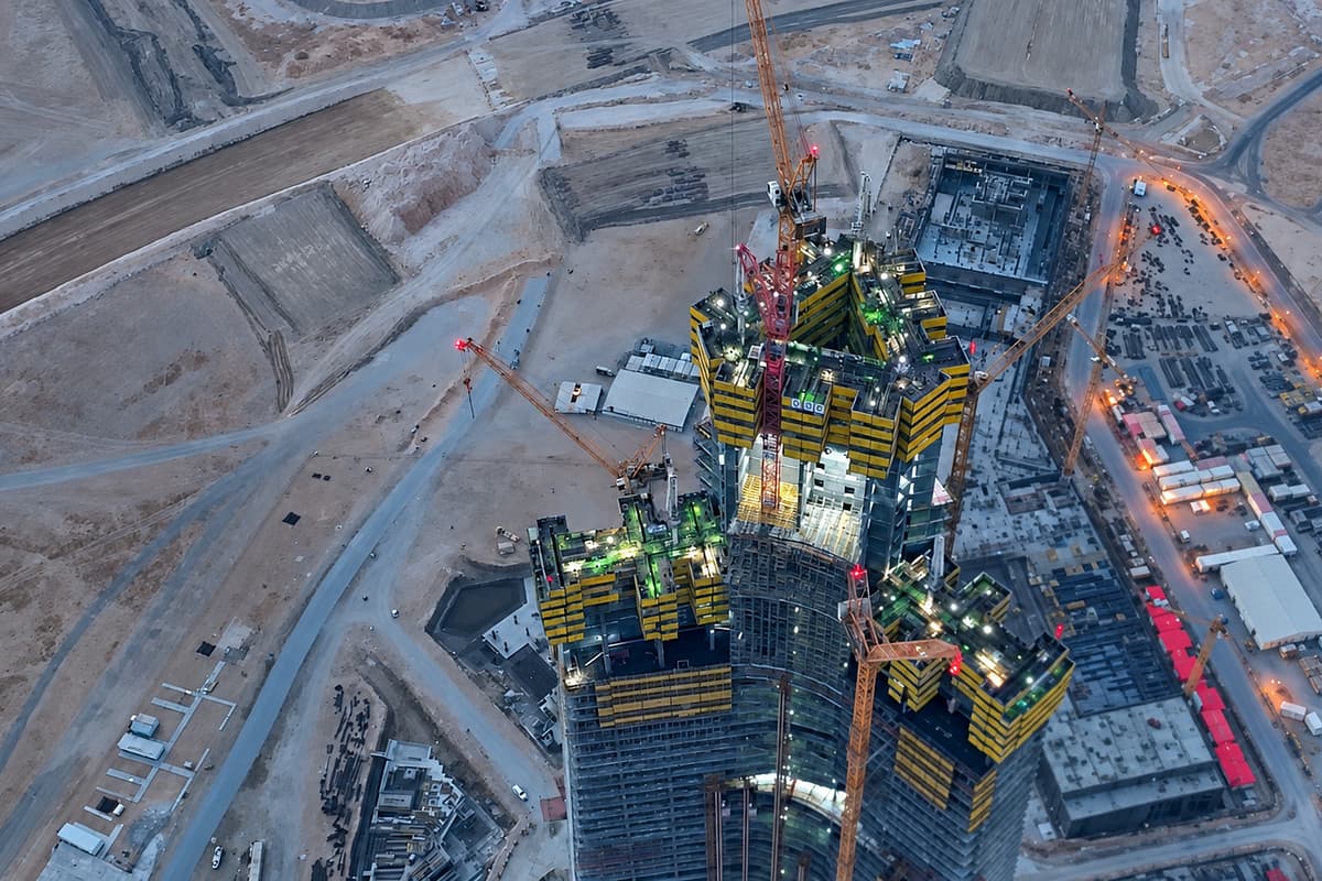 Aerial view of a large high-rise building under construction, surrounded by multiple tower cranes, construction equipment, materials, and access roads in a desert landscape