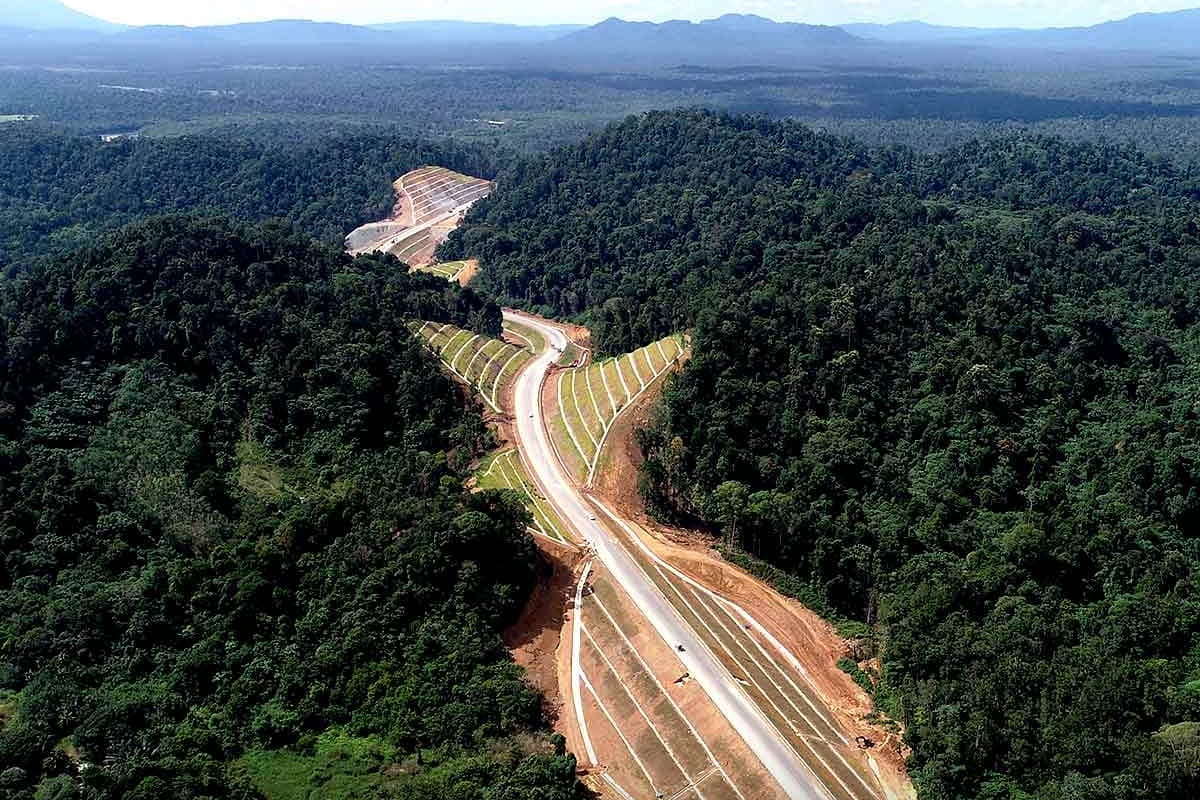 Aerial view of a newly built highway winding through dense tropical forest, with construction slopes and distant blue mountains under a bright sky.