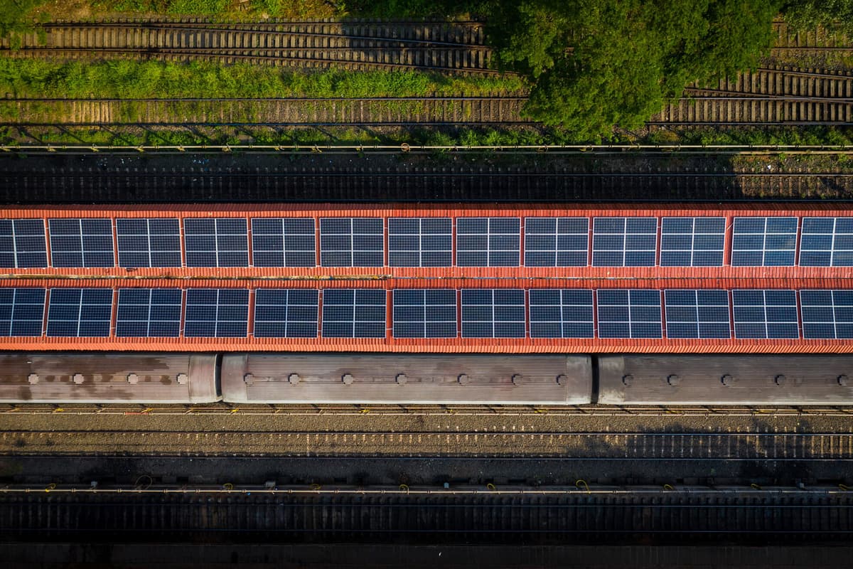 Aerial view of parallel railway tracks and a long red-and-blue covered bridge, bordered by green vegetation and industrial infrastructure.