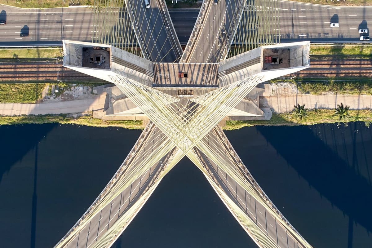 Aerial view of a cable-stayed bridge crossing a dark river in an X-shaped pattern, with highways and rail lines above; striking geometry.