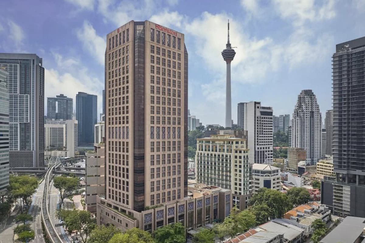 Kuala Lumpur skyline with KL Tower rising behind a historic office building, modern high-rises, busy roads, greenery, and bright blue skies.