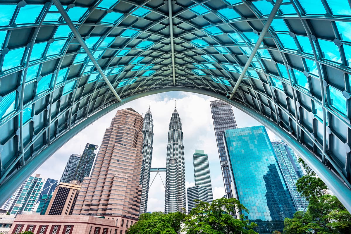 A low-angle view of the Petronas Twin Towers and surrounding skyscrapers in Kuala Lumpur, framed by the arch of a modern walkway with a blue-tinted glass roof.