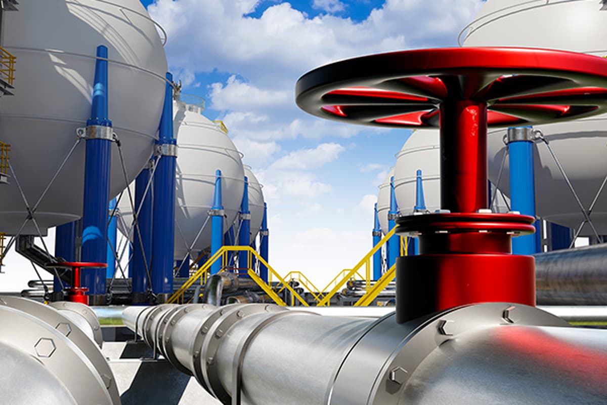 Close-up of a red industrial valve on a silver pipeline, with blue-and-white storage tanks, yellow stairs, and a bright cloudy sky behind.