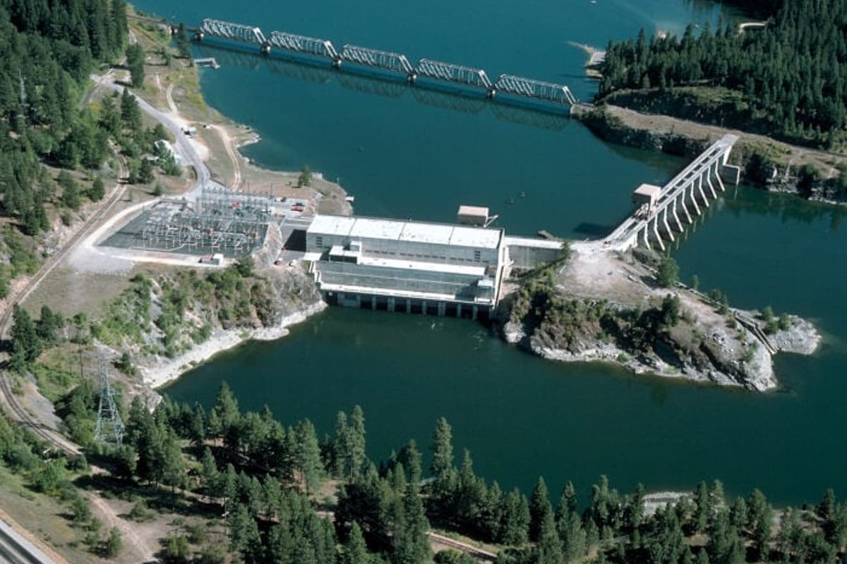 Aerial view of a hydroelectric dam and spillways spanning a forested reservoir, with roads, power lines, and green mountainside surroundings.
