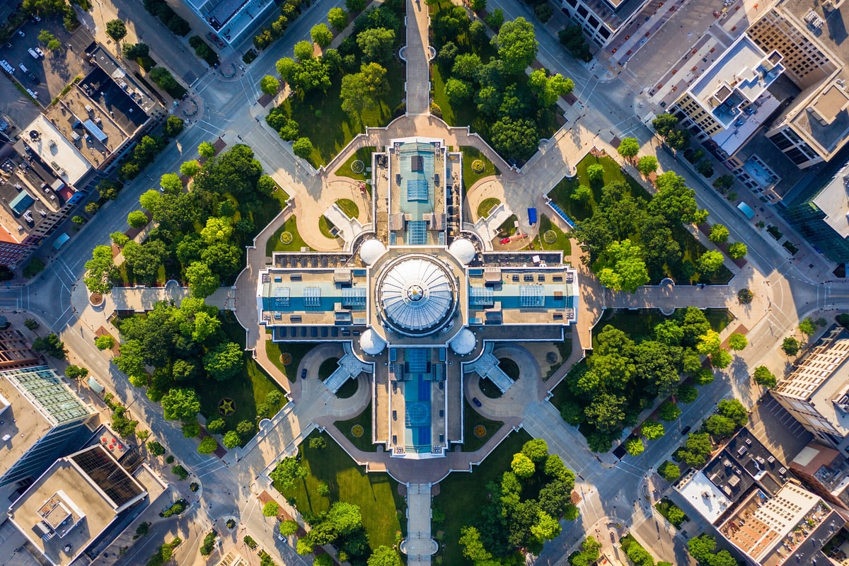 Aerial view of a grand domed building with symmetrical wings, surrounded by tree-lined paths, streets, and dense city blocks.