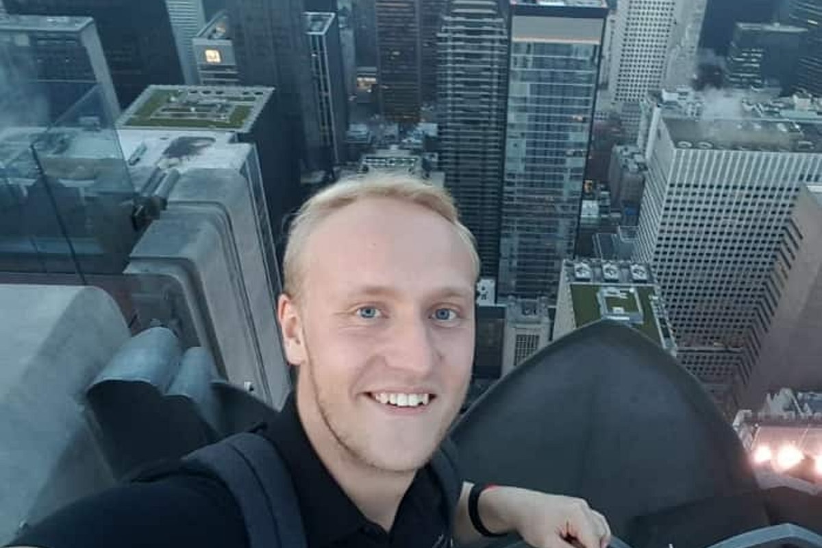 Smiling man in black shirt leans over fence with cityscape in background