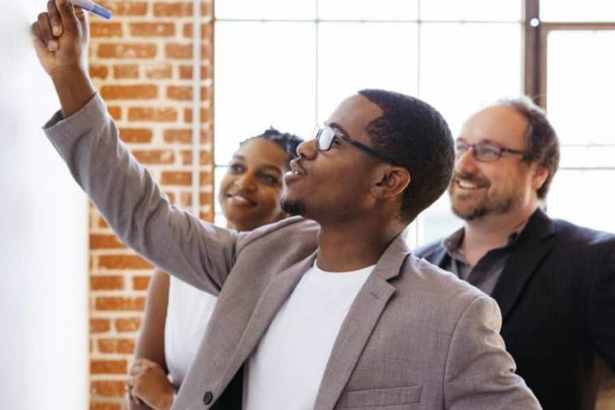Man points upward with pen in hand while man and woman in background look upward smiling