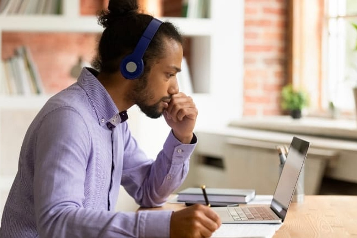 Man with headphones holds pen while working at laptop with office setting in the background