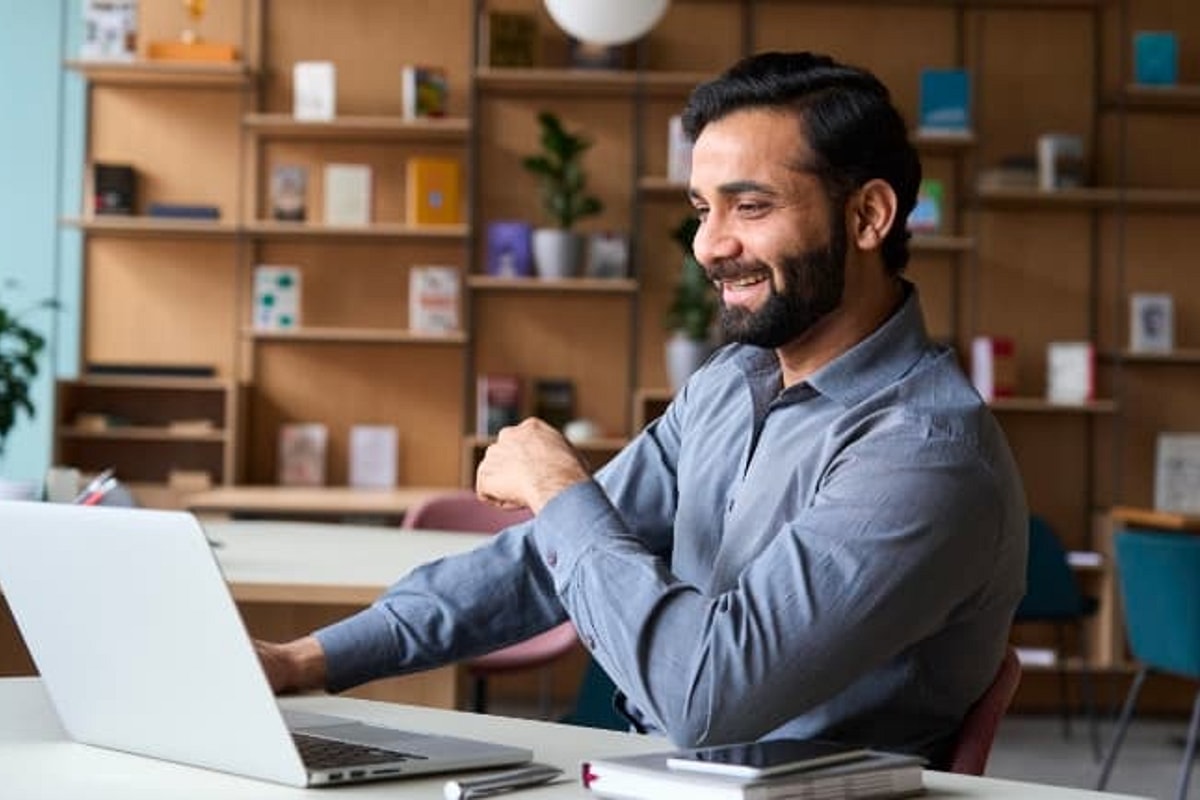 Man in blue shirt smiles while looking at laptop on desk with office setting in background