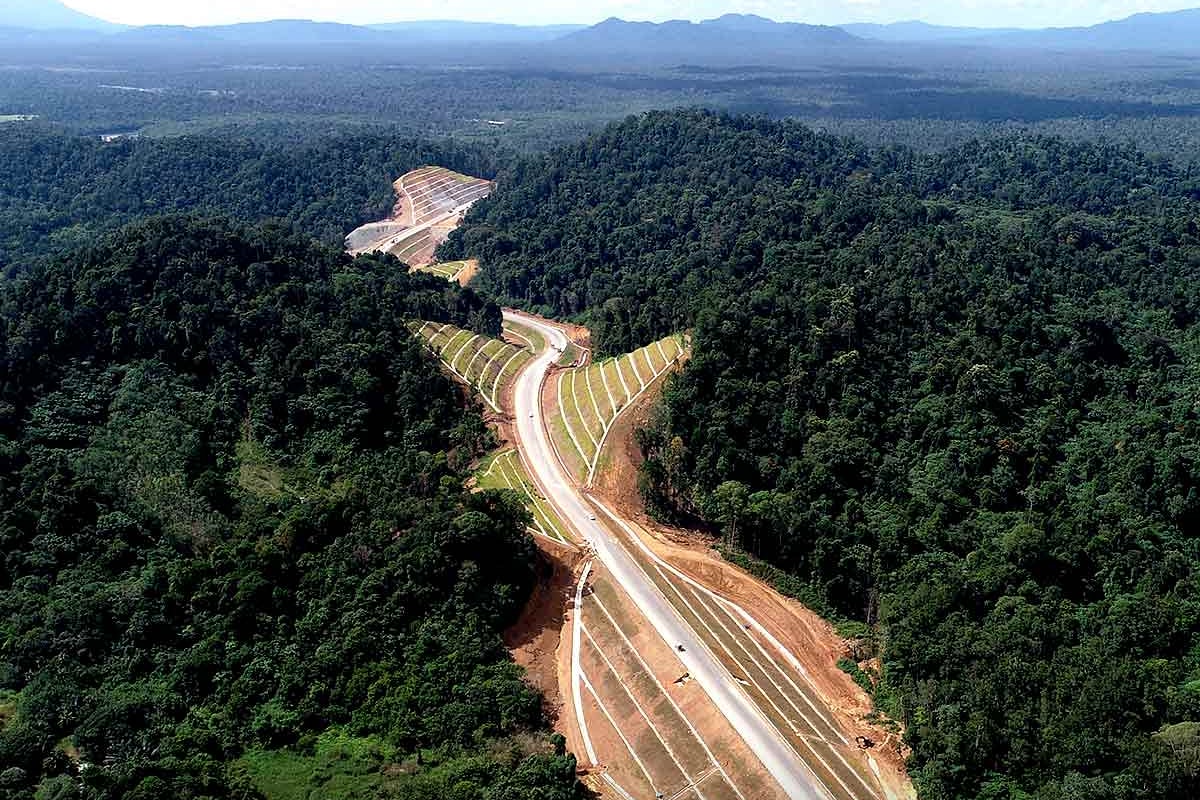 Aerial view of a newly built highway winding through dense tropical forest, with construction slopes and distant blue mountains under a bright sky.