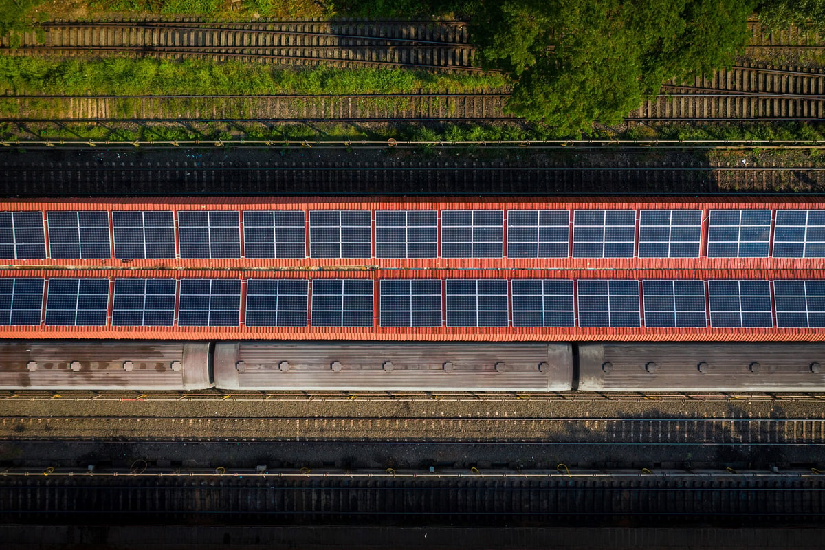 Aerial view of parallel railway tracks and a long red-and-blue covered bridge, bordered by green vegetation and industrial infrastructure.