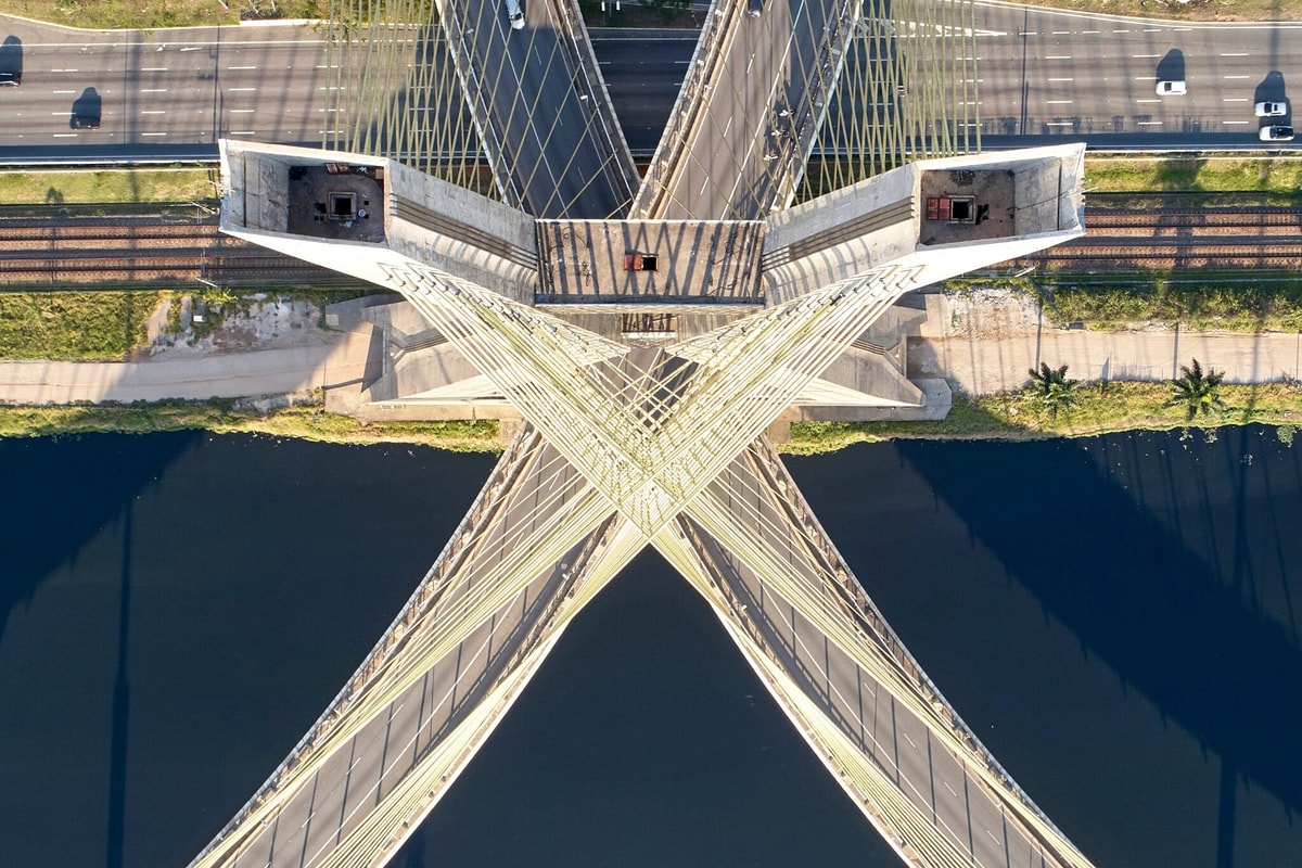 Aerial view of a cable-stayed bridge crossing a dark river in an X-shaped pattern, with highways and rail lines above; striking geometry.
