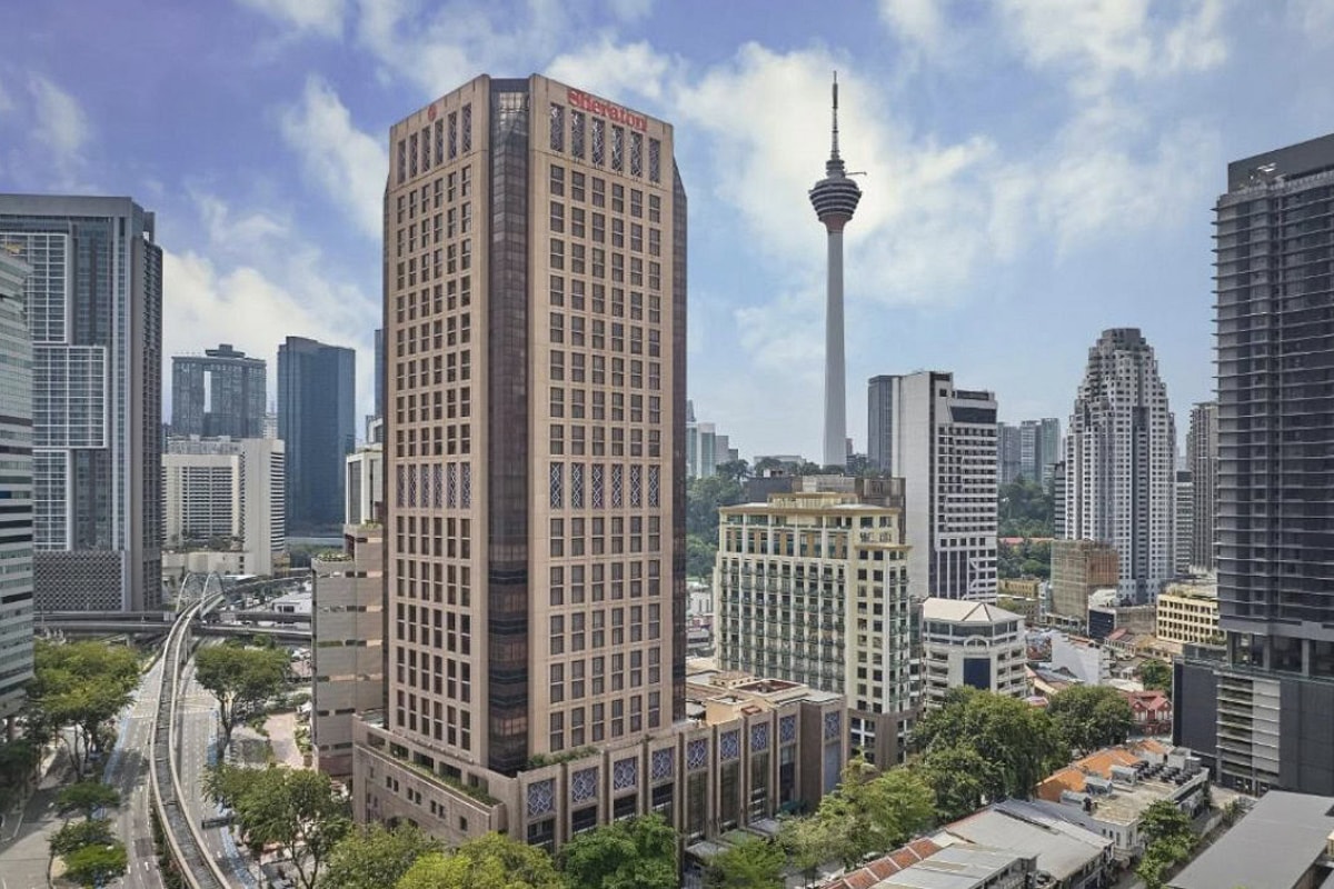 Kuala Lumpur skyline with KL Tower rising behind a historic office building, modern high-rises, busy roads, greenery, and bright blue skies.