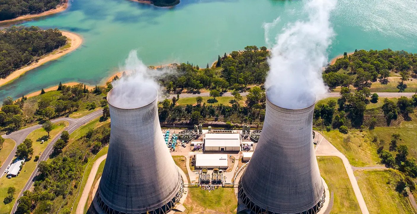 Aerial view of a conventional power plant with twin concrete cooling towers emitting steam, nestled near a scenic water reservoir and illustrating civil engineering and power generation infrastructure.