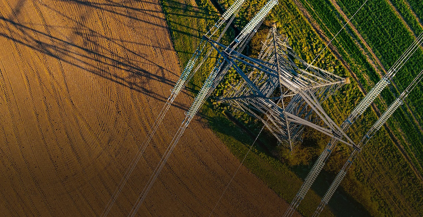 Direct top-down aerial view of a steel lattice transmission tower and high-voltage power lines traversing a rural landscape, illustrating grid connectivity, bulk power transmission, and energy sector reliability.