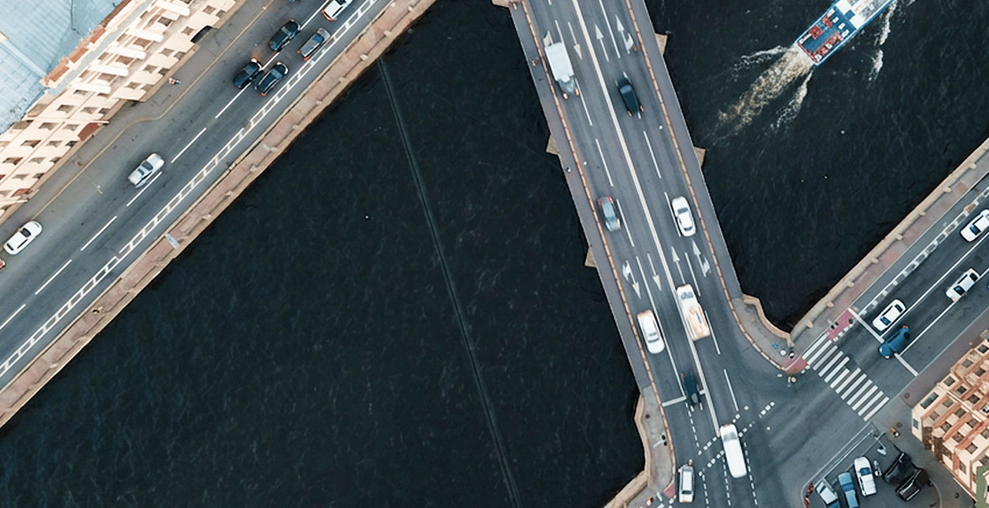 An aerial view of an active multi-lane roadway bridge crossing a river with a passenger boat traveling below, illustrating civil engineering, public transportation, and connected urban infrastructure.
