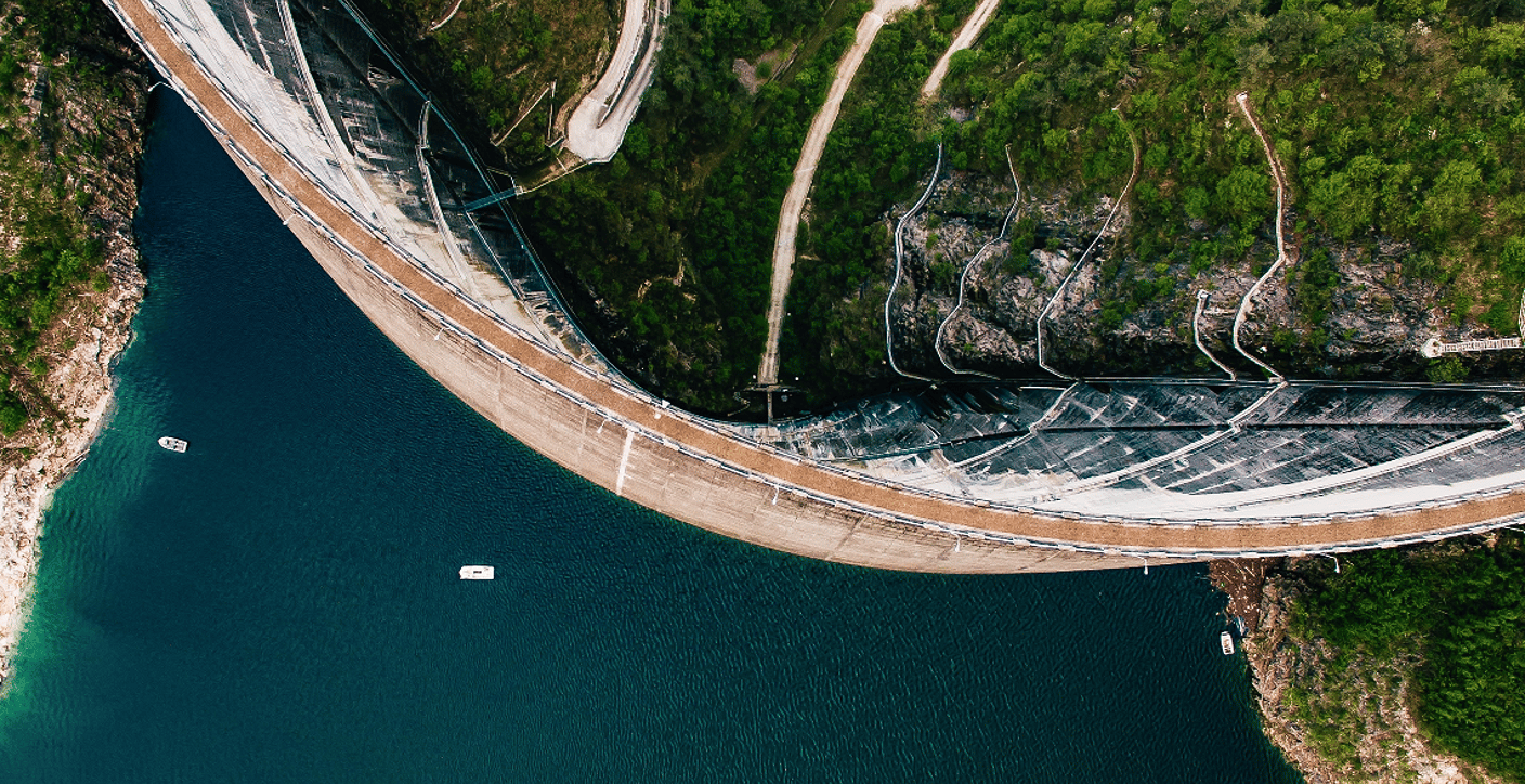 A high-altitude aerial view of a curved concrete arch dam impounding a deep blue reservoir, showcasing large-scale water storage, resource conservation, and civil engineering.
