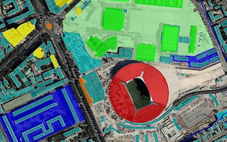 A top-down GIS map view in OpenCities Map featuring a stadium with a red circular roof, surrounded by green, blue, and yellow color-coded building plots for urban planning.