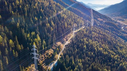 Aerial view of overhead power lines running through a forested mountain landscape under a clear sky, showcasing impressive power line engineering with distant valleys visible.