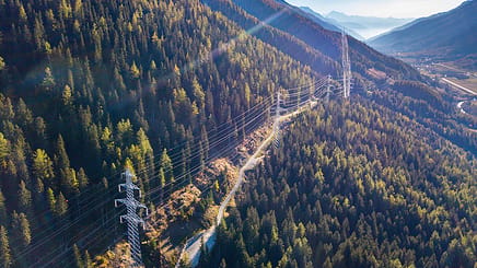 Aerial view of overhead power lines running through a forested mountain landscape under a clear sky, showcasing impressive power line engineering with distant valleys visible.