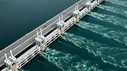 An aerial view of a large-scale storm surge barrier with a road running along its top. The structure spans a body of water, with calm, dark water on one side and turbulent, churning turquoise water on the other, indicating a powerful flow through its gates.