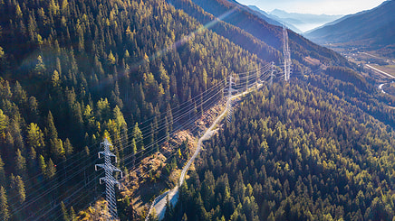 Aerial view of overhead power lines running through a forested mountain landscape under a clear sky, showcasing impressive power line engineering with distant valleys visible.