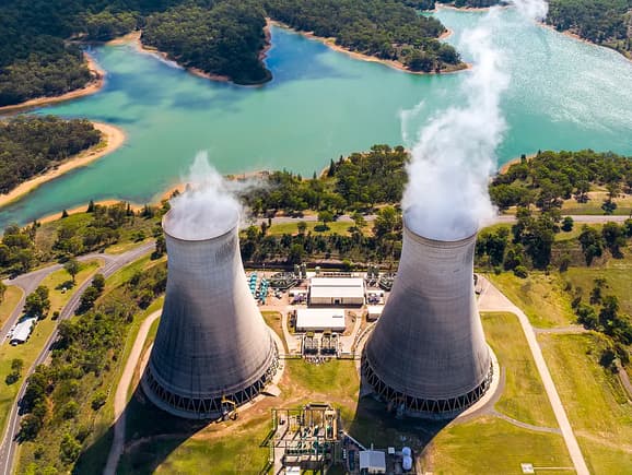 Aerial view of a conventional power plant with twin concrete cooling towers emitting steam, nestled near a scenic water reservoir and illustrating civil engineering and power generation infrastructure.