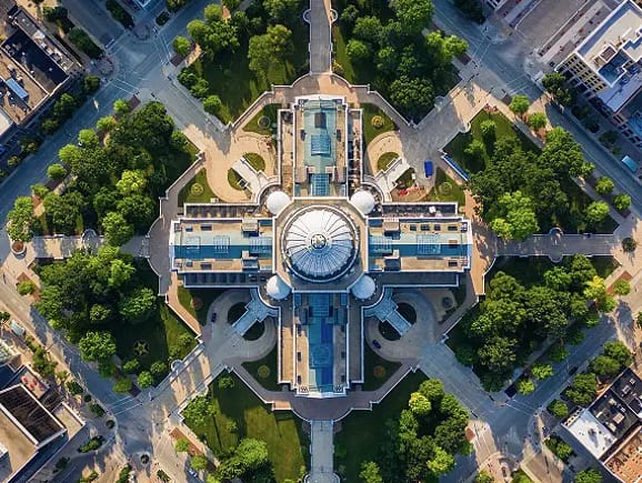 A symmetrical high-angle aerial view of a grand dome-topped capitol building surrounded by manicured lawns, circular plazas, and radial pathways, representing civic architecture and government infrastructure.