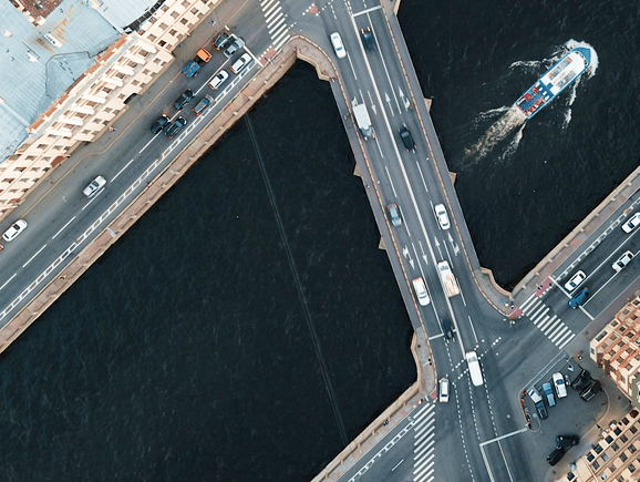 An aerial view of an active multi-lane roadway bridge crossing a river with a passenger boat traveling below, illustrating civil engineering, public transportation, and connected urban infrastructure.