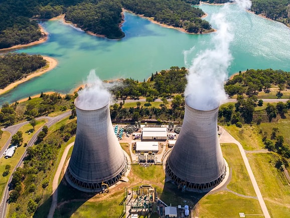 Aerial view of a conventional power plant with twin concrete cooling towers emitting steam, nestled near a scenic water reservoir and illustrating civil engineering and power generation infrastructure.
