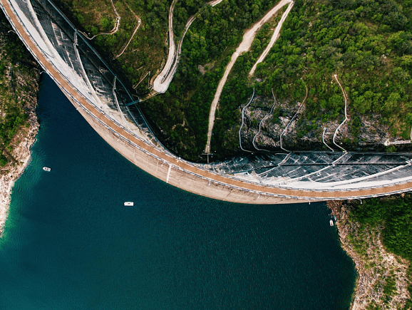 A high-altitude aerial view of a curved concrete arch dam impounding a deep blue reservoir, showcasing large-scale water storage, resource conservation, and civil engineering.