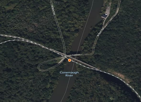 AssetWise Inspections GIS mapping interface showing a circular orange asset location marker positioned on a bridge crossing the Conemaugh River on the W Penn Trail, surrounded by dense forest.