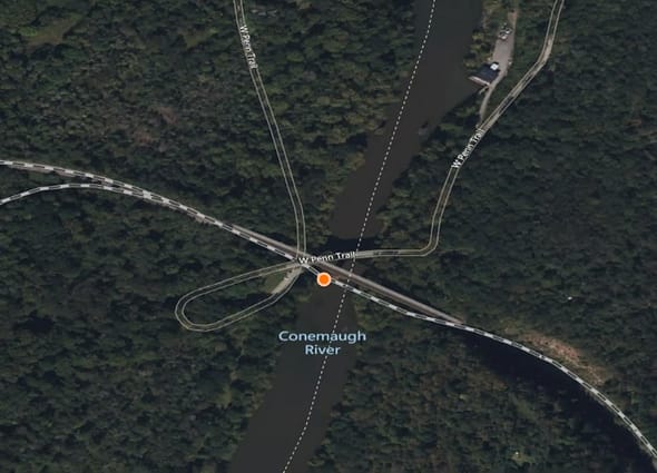 AssetWise Inspections GIS mapping interface showing a circular orange asset location marker positioned on a bridge crossing the Conemaugh River on the W Penn Trail, surrounded by dense forest.