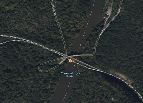 AssetWise Inspections GIS mapping interface showing a circular orange asset location marker positioned on a bridge crossing the Conemaugh River on the W Penn Trail, surrounded by dense forest.