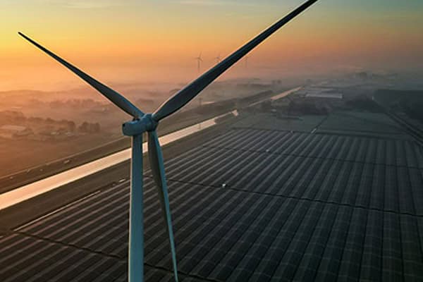A towering wind turbine rises above vast solar-panel fields at sunrise, its blades silhouetted against a hazy orange sky, evoking clean energy.