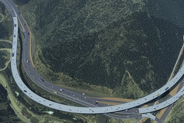 Aerial view of a sweeping highway interchange curving around a forested hillside, with cars and trucks traveling through the green landscape.
