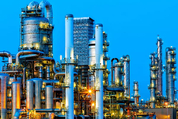 Industrial oil refinery with towering distillation columns, pipes, and tanks glowing under orange lights against a vivid blue evening sky.