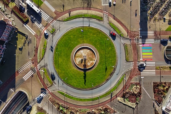 An aerial top-down view of an urban roundabout featuring a green central island, structured traffic lanes, red bicycle lanes, and pedestrian crosswalks, highlighting smart city design and urban mobility planning.