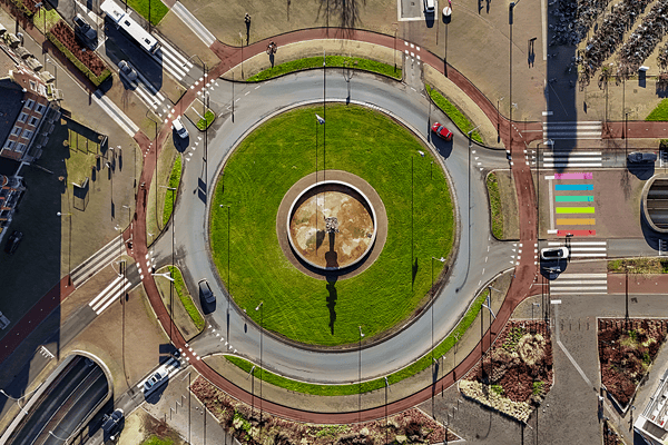 An aerial top-down view of an urban roundabout featuring a green central island, structured traffic lanes, red bicycle lanes, and pedestrian crosswalks, highlighting smart city design and urban mobility planning.