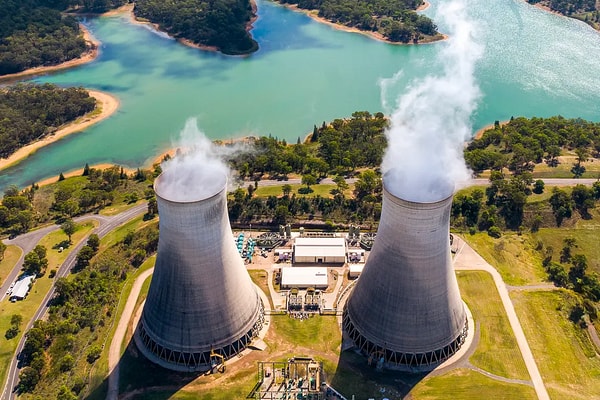 Aerial view of a conventional power plant with twin concrete cooling towers emitting steam, nestled near a scenic water reservoir and illustrating civil engineering and power generation infrastructure.