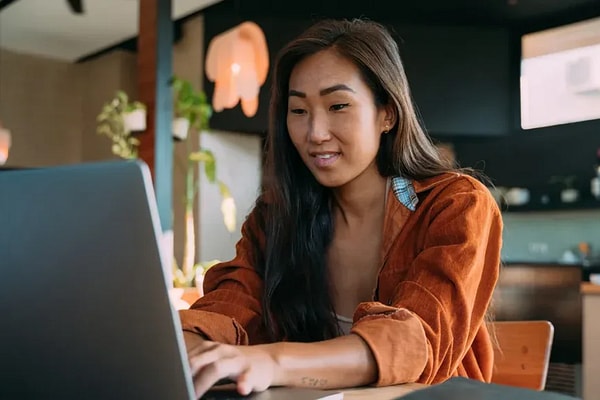 Woman with long hair and orange shirt works at laptop with office setting in background