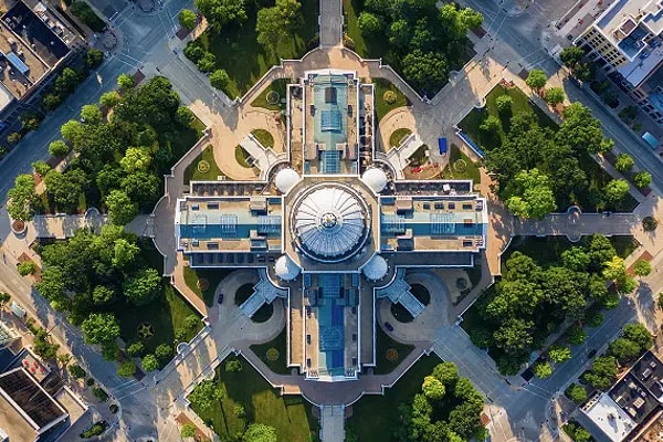 A symmetrical high-angle aerial view of a grand dome-topped capitol building surrounded by manicured lawns, circular plazas, and radial pathways, representing civic architecture and government infrastructure.