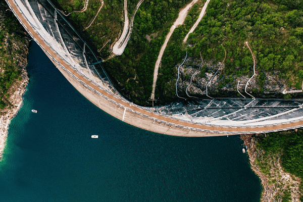 A high-altitude aerial view of a curved concrete arch dam impounding a deep blue reservoir, showcasing large-scale water storage, resource conservation, and civil engineering.