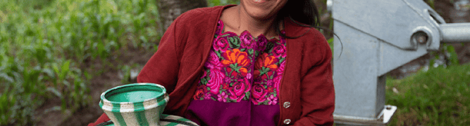 A smiling woman in the community of Panyey, Guatemala, wearing traditional clothing, holds a water jug while standing next to a newly installed water pump that brings clean, running water to her village.