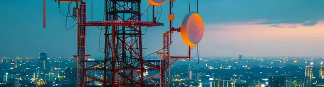 A telecommunications tower with multiple antennas and dishes overlooks an illuminated cityscape at dusk.