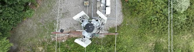 A top-down aerial view of a communications tower and power lines over a lush green forest.