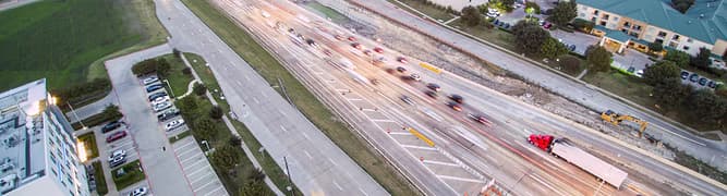 An aerial view of a busy highway in Plano, Texas, with blurred evening traffic flowing past the commercial district.