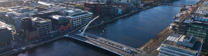 Aerial view of a modern bridge spanning a river in a city, with surrounding buildings and roads visible under a clear sky.